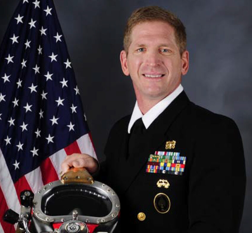 Commander Joseph Dituri in U.S. Navy dress blues, one hand resting on a brass diving helmet, an American flag behind him