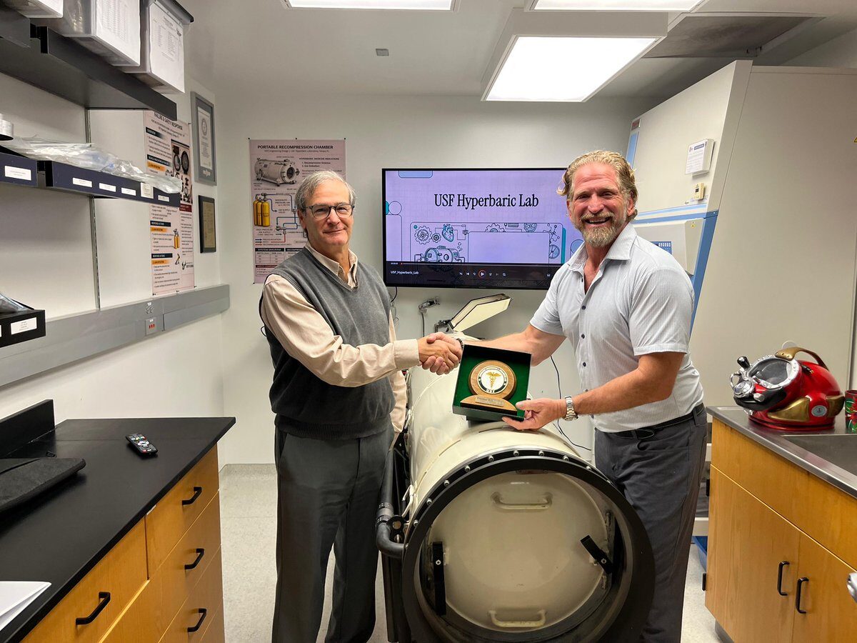 Joseph Dituri shaking hands with a colleague in the USF Hyperbaric Laboratory, the two of them holding an open presentation case between them, a horizontal recompression chamber filling the foreground and a screen reading USF Hyperbaric Lab on the wall behind