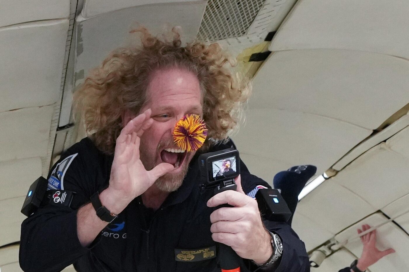 Joseph Dituri laughing in zero gravity aboard a parabolic flight, his hair floating around his head, a camera in one hand and a foam ball drifting in front of his face