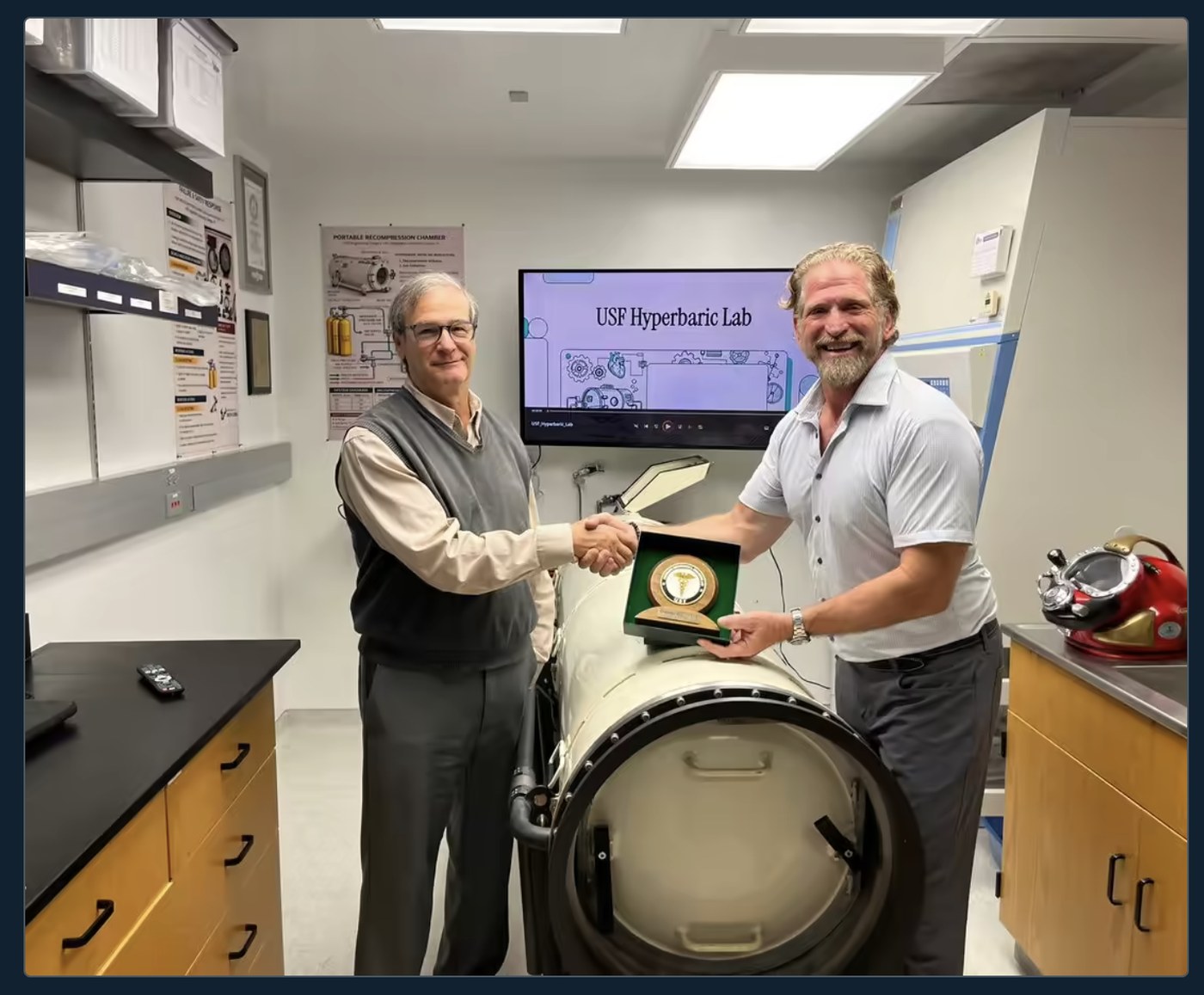 Joseph Dituri and a colleague shaking hands beside a portable recompression chamber in the USF Hyperbaric Lab, a plaque between them and a presentation on the screen behind